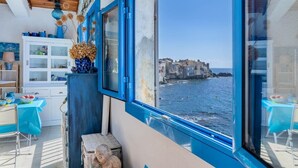 A room with a view of a coastal town, a blue-framed window, a wicker chair, and a small table with a blue tablecloth. - Casetta Ricci - Les Azedarachs II (Brando)