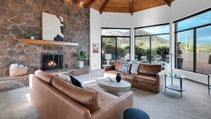 A spacious living room with a stone fireplace, leather sofas, and large windows offering a view of the desert landscape. - Catalina Dreamin' Luxe Foothills Oasis w/Pool! (Tucson)