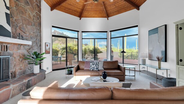 A spacious living area with a stone fireplace, leather sofas, and large windows offering a view of cacti and mountains. - Catalina Dreamin' Luxe Foothills Oasis w/Pool! (Tucson)