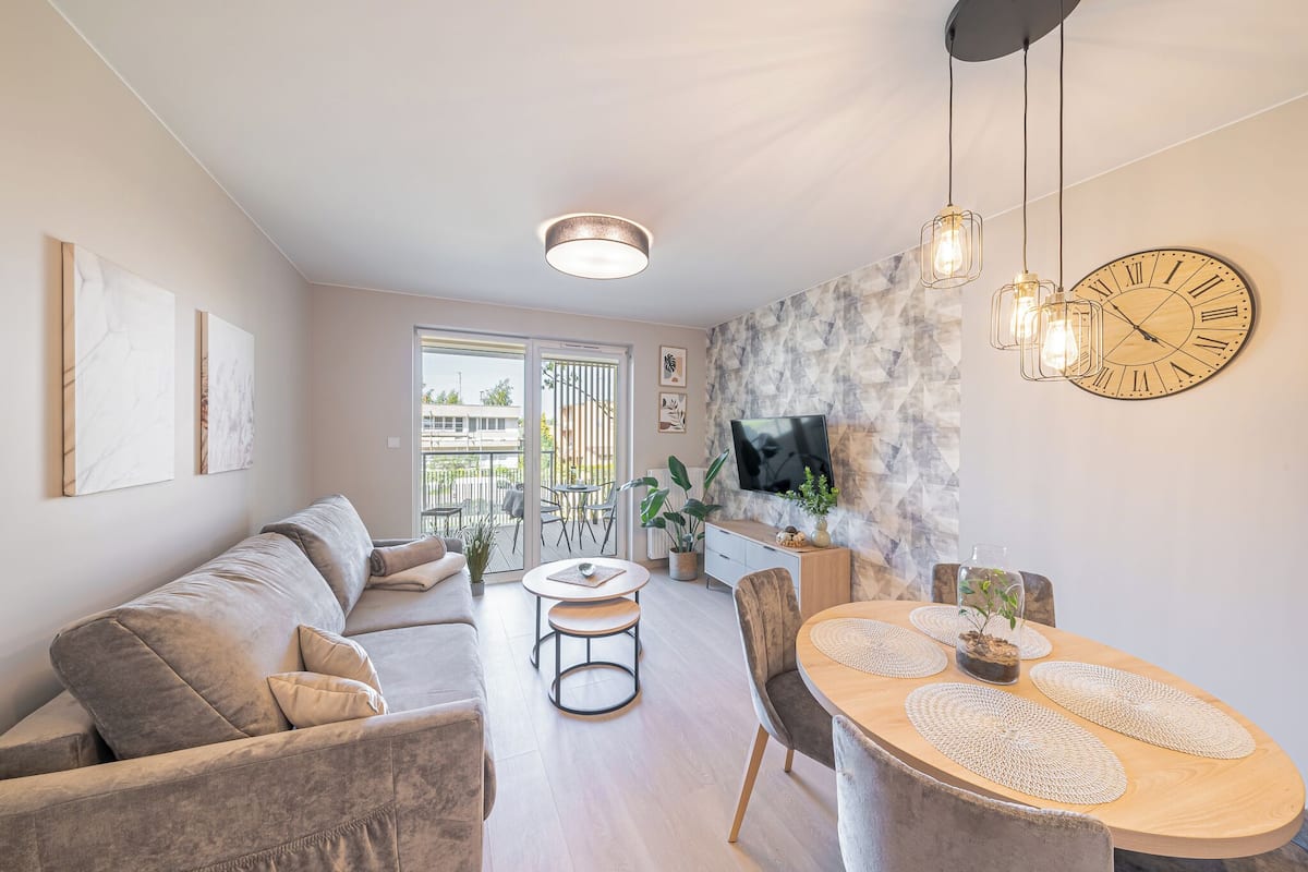 Ein modernes Wohnzimmer mit einer Couch, einem Esstisch, Stühlen, einem an einer Steinwand montierten Fernseher und einem Balkon mit Gartenmöbeln.