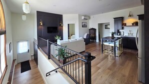 Un amplio espacio de estar con techo alto, una escalera con barandilla, una cocina con armarios oscuros y un comedor con mesa y sillas.