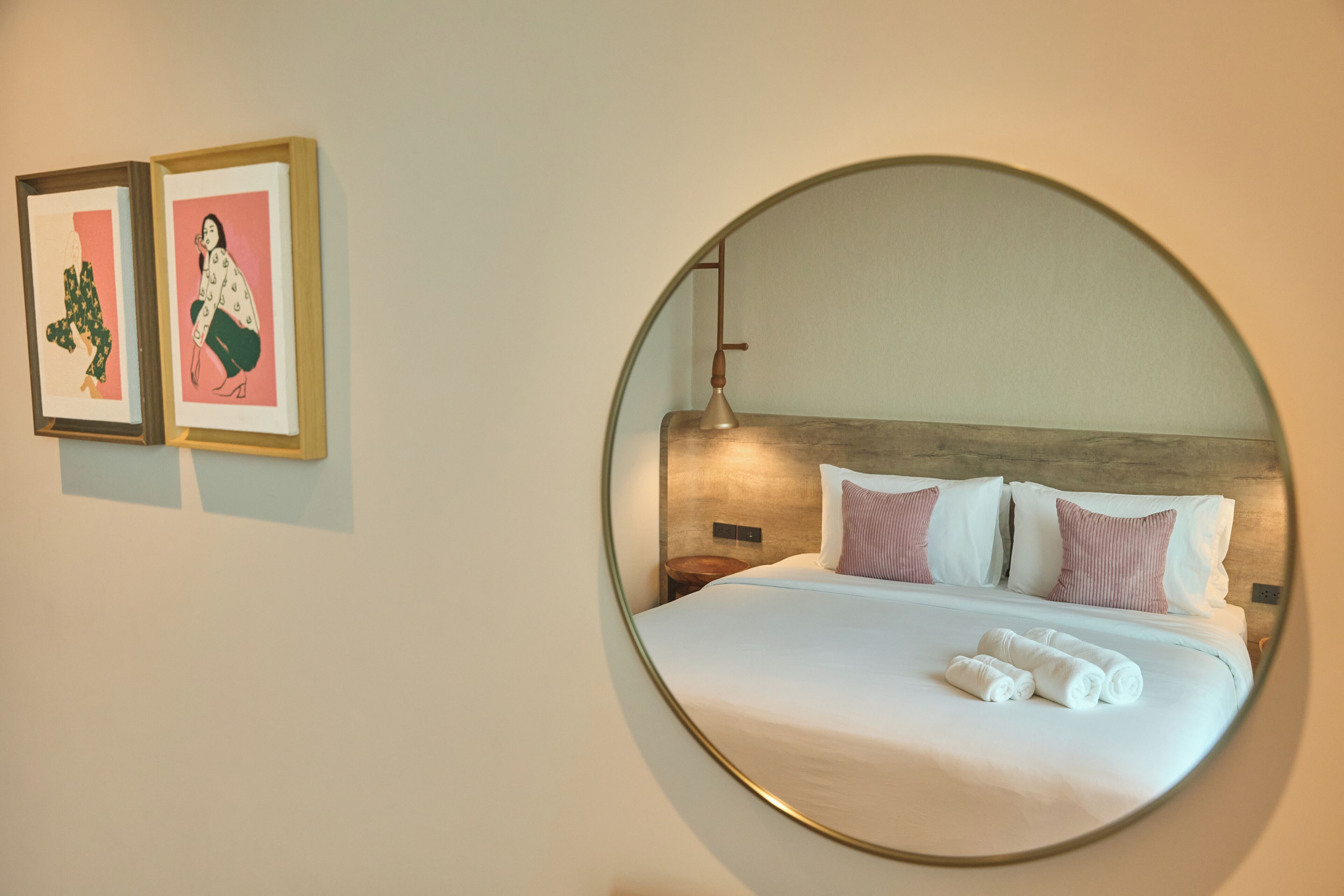 Habitación de hotel con un espejo grande y redondo, una cama bien hecha con almohadas y dos obras de arte enmarcadas en la pared.