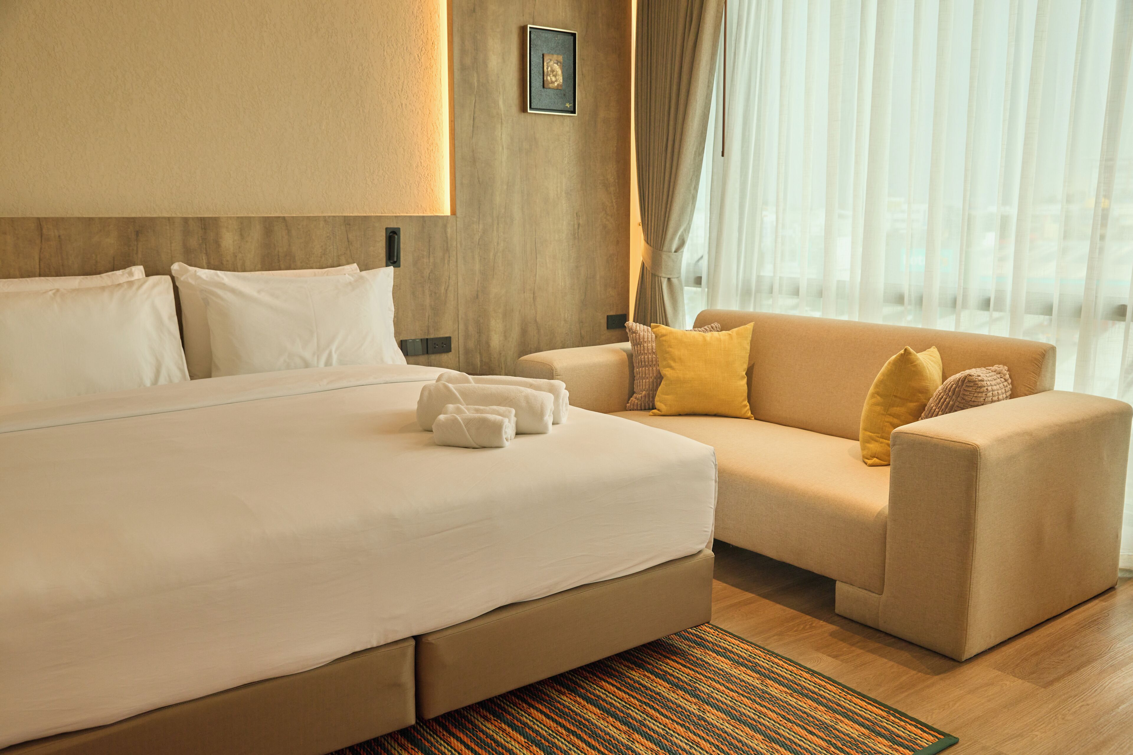 Quarto de hotel com cama grande, sofá e vista da cidade através de cortinas translúcidas.
