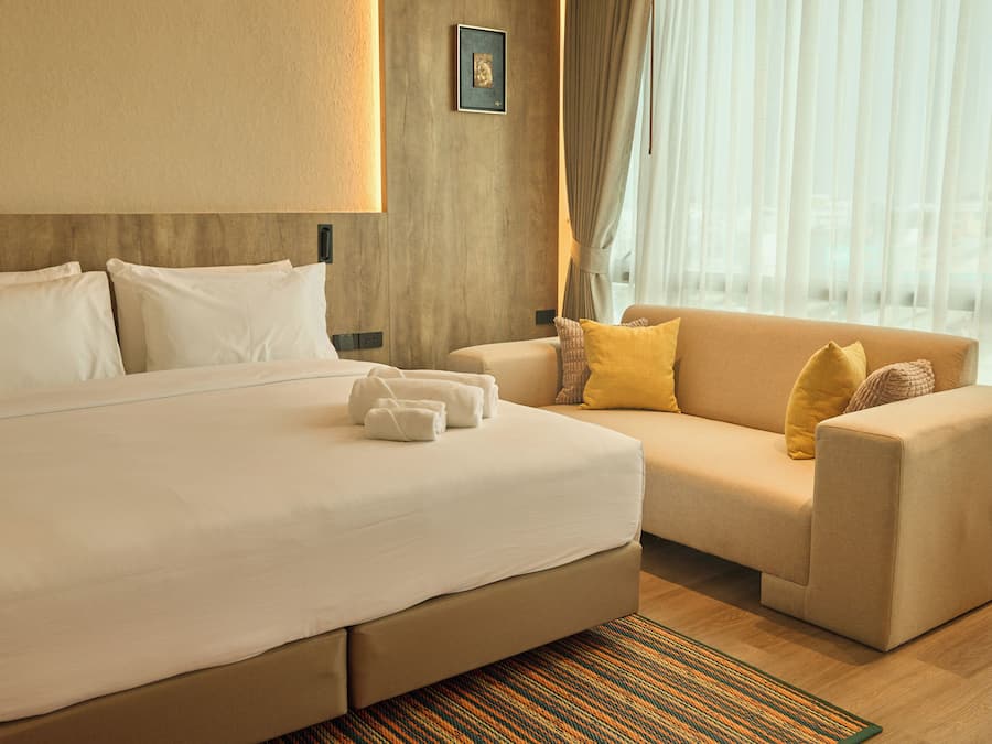 Quarto de hotel com cama grande, sofá e vista da cidade através de cortinas translúcidas.