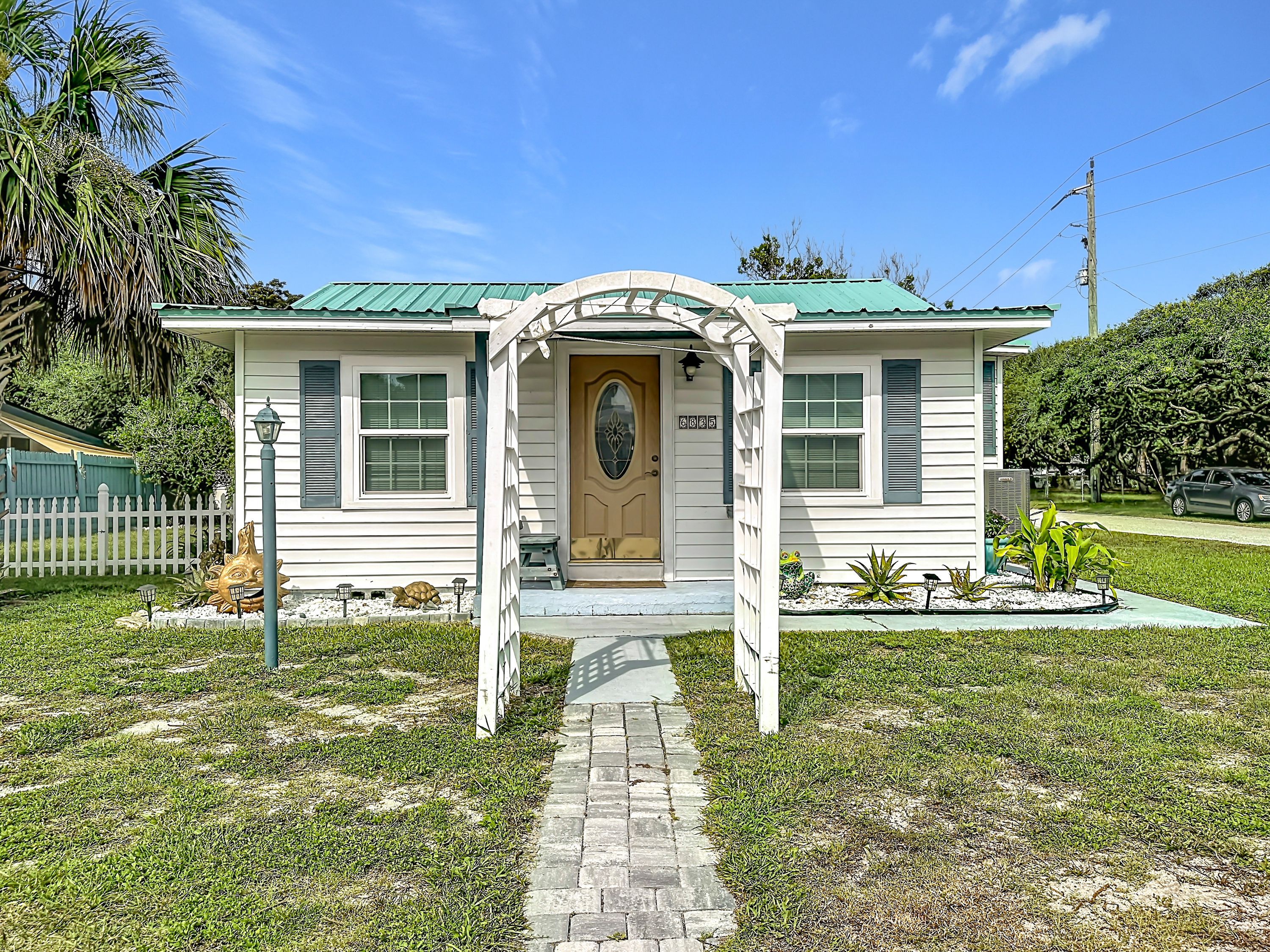 A small, single-story house with a green roof, a front porch, and a pathway leading to the entrance.