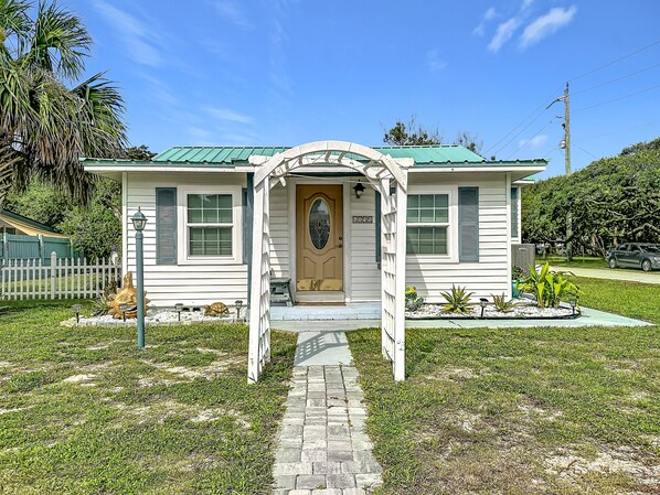 A small, single-story house with a green roof, a front porch, and a pathway leading to the entrance. - Crescent Beach Cottage | Dog-Friendly (St. Augustine)