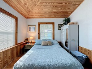 A bedroom with a bed, a nightstand, a lamp, a dresser, and a window with blinds. - Crescent Beach Cottage | Dog-Friendly (St. Augustine)