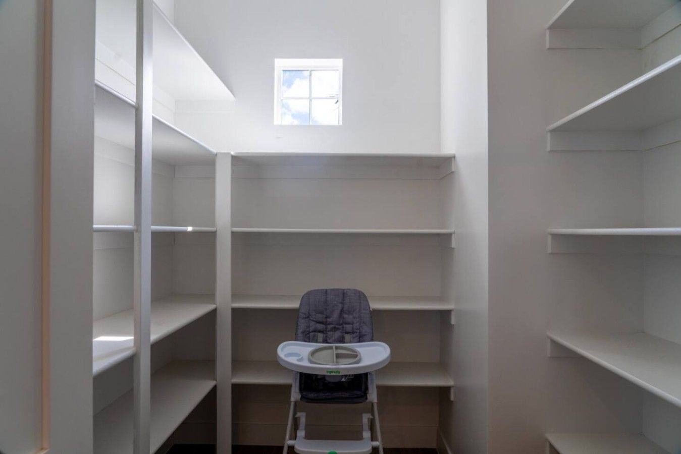 A narrow, empty walk-in closet with white shelving and a small window at the top.
