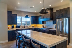 Una cocina moderna con gabinetes oscuros, una isla grande con taburetes de bar, electrodomésticos de acero inoxidable e iluminación colgante.
