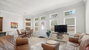 A spacious living room with a large brown leather sofa, a wooden coffee table, and a television on a wooden stand. There is a dining area with a wooden table and chairs, and a potted plant near the window.