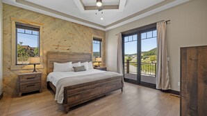 Ein Schlafzimmer mit einem großen Bett, einem Holzkopfteil, zwei Nachttischen mit Lampen, einem Deckenventilator und Blick ins Freie durch eine Glastür.