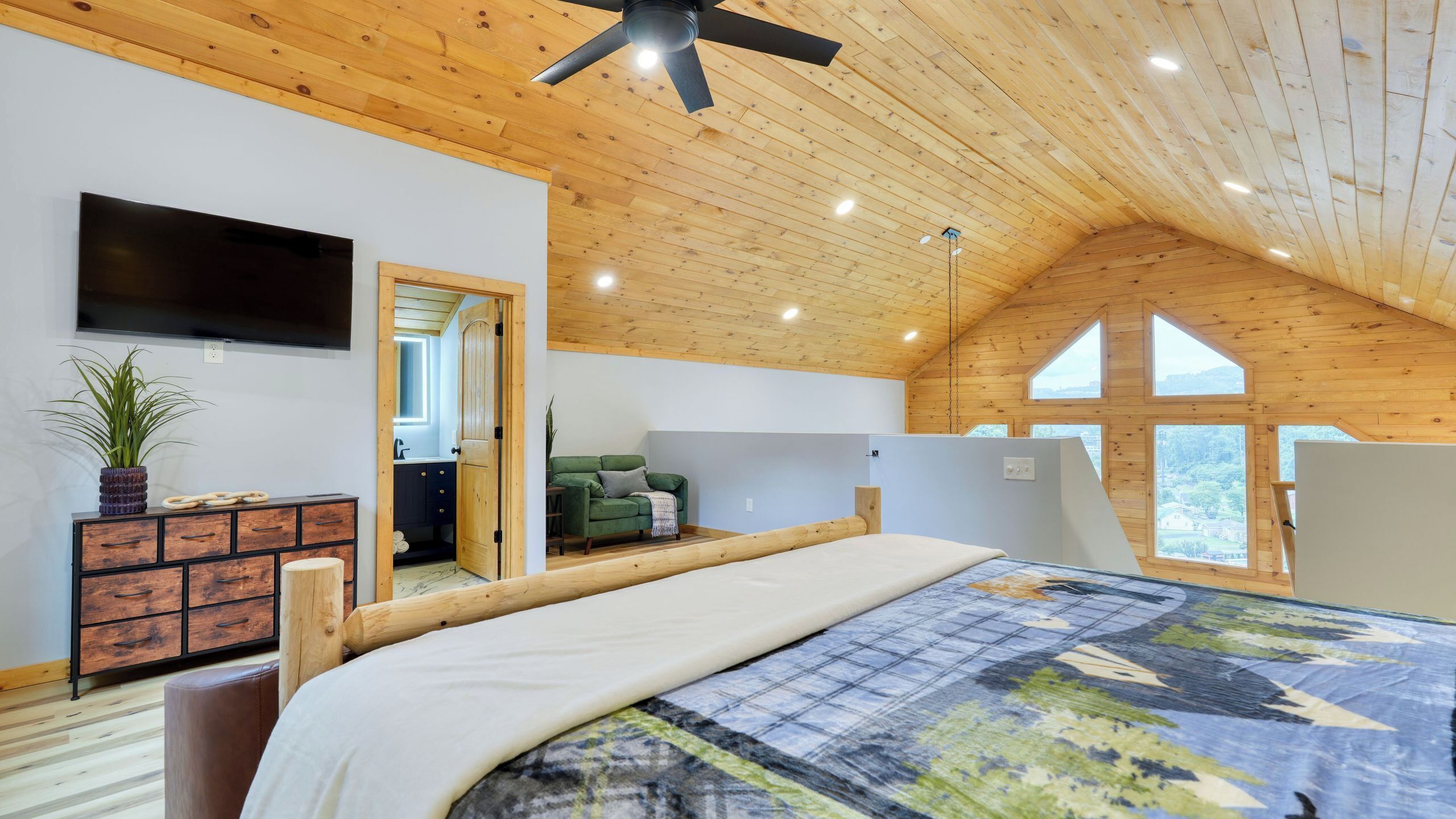 Un dormitorio con techo de madera, una cama con colcha estampada, un sofá verde, un televisor y vistas por una ventana.