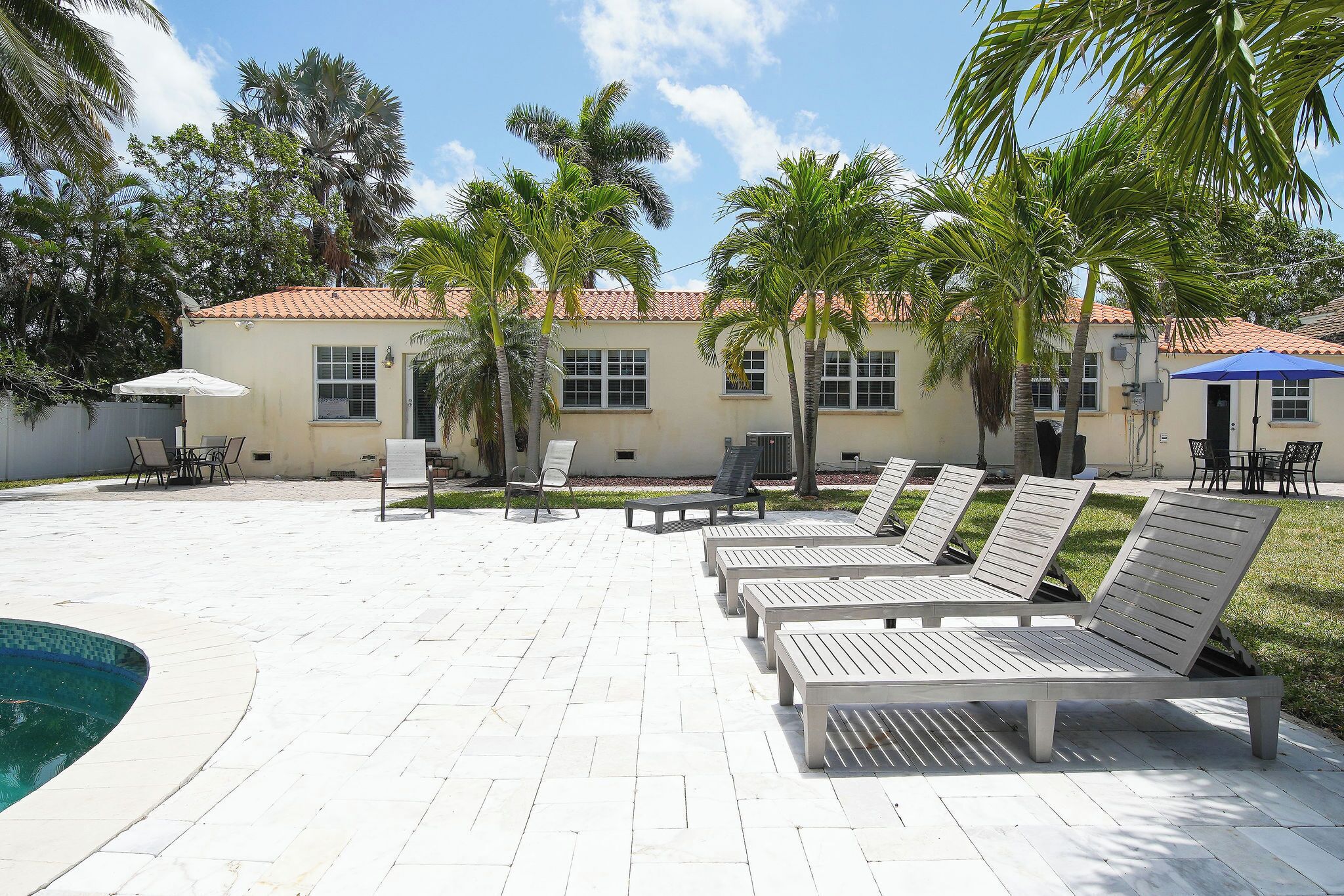 A poolside area with lounge chairs and umbrellas, palm trees, and a building with a tiled roof.