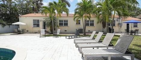 A poolside area with lounge chairs and umbrellas, palm trees, and a building with a tiled roof.