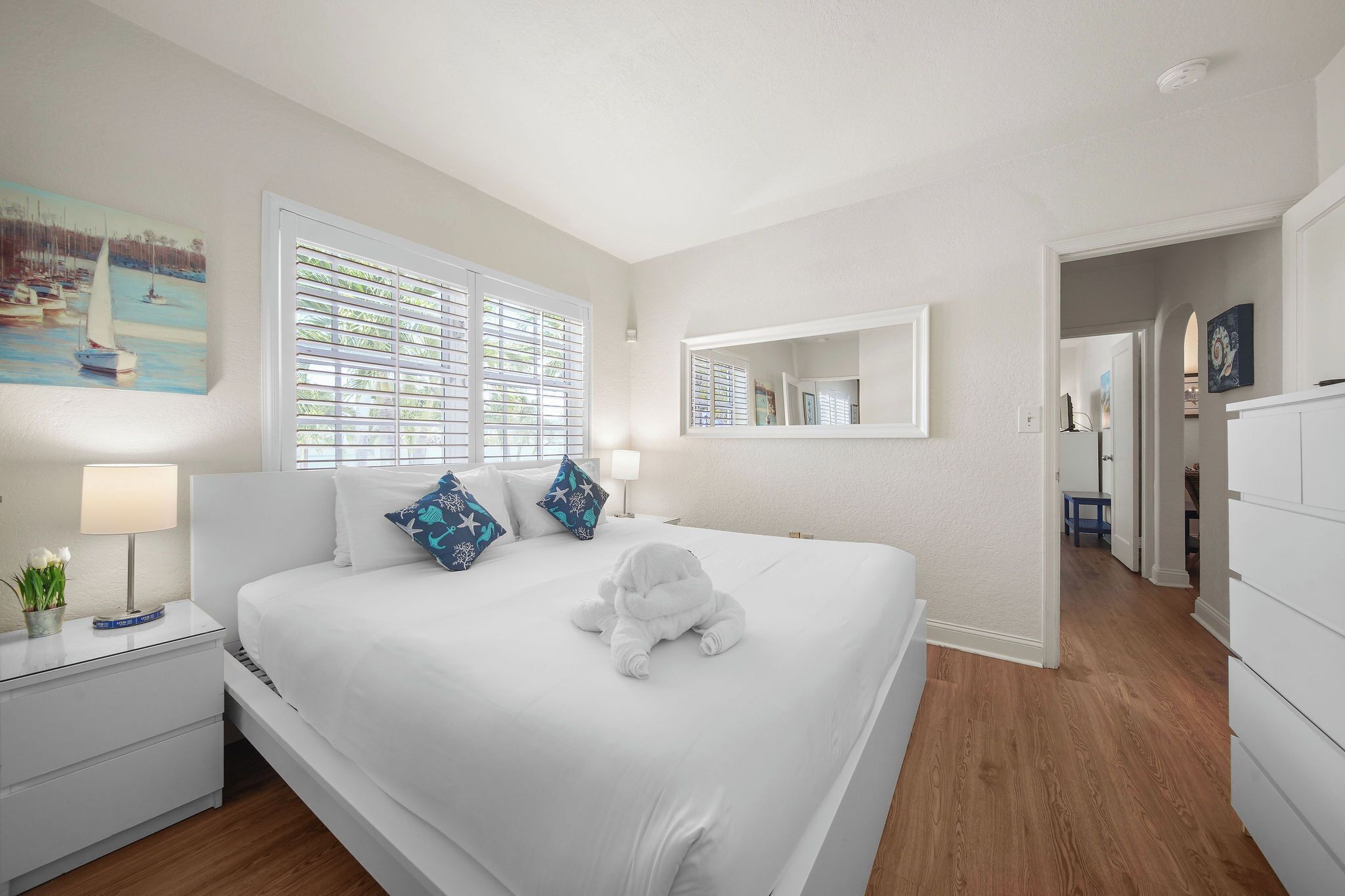 A neatly made bed with a white comforter and pillows, a bedside table with a lamp and a small plant, a large window with blinds, and a mirror on the wall.