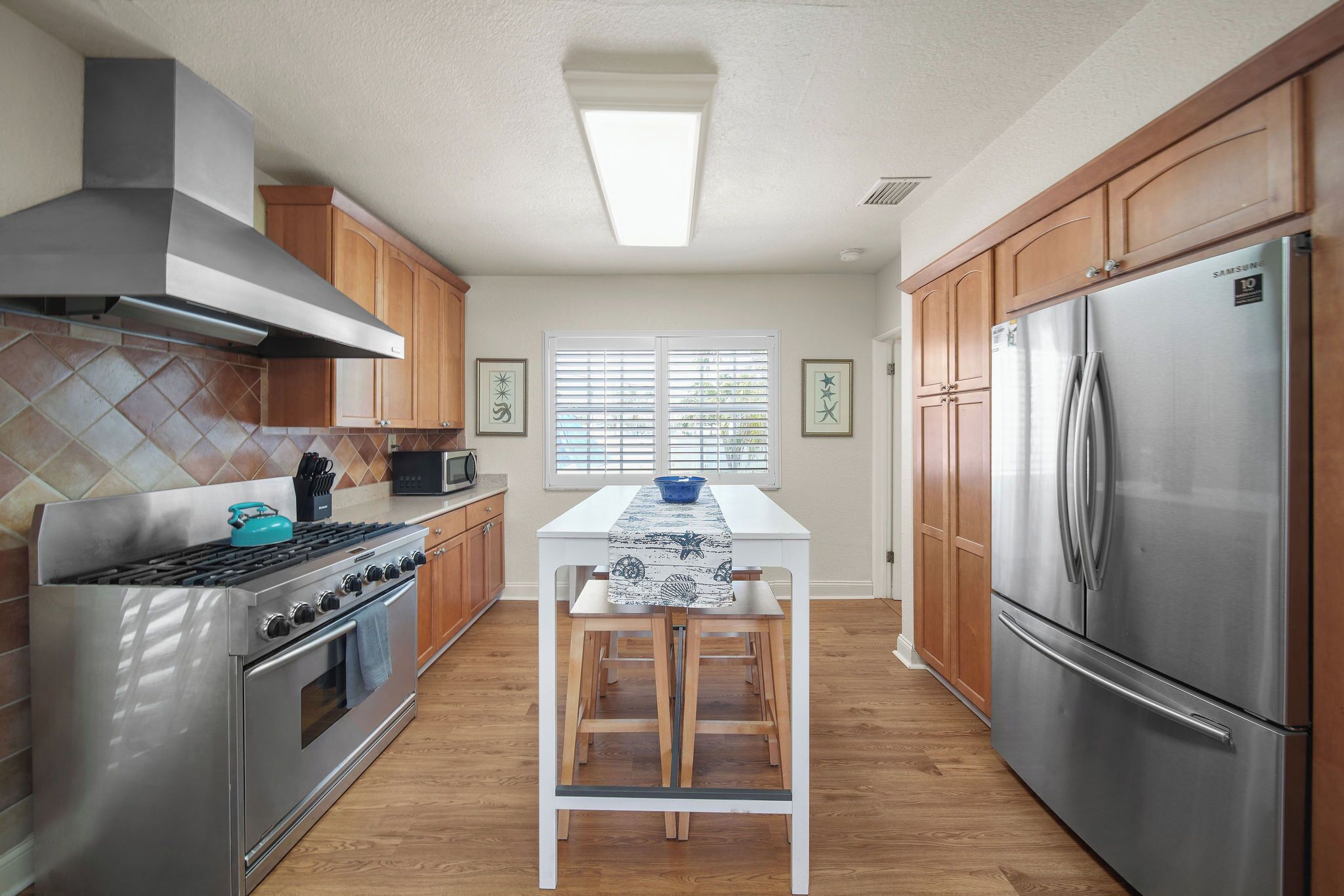 A modern kitchen with wooden cabinets, a stainless steel refrigerator, a gas stove, and a window with blinds.