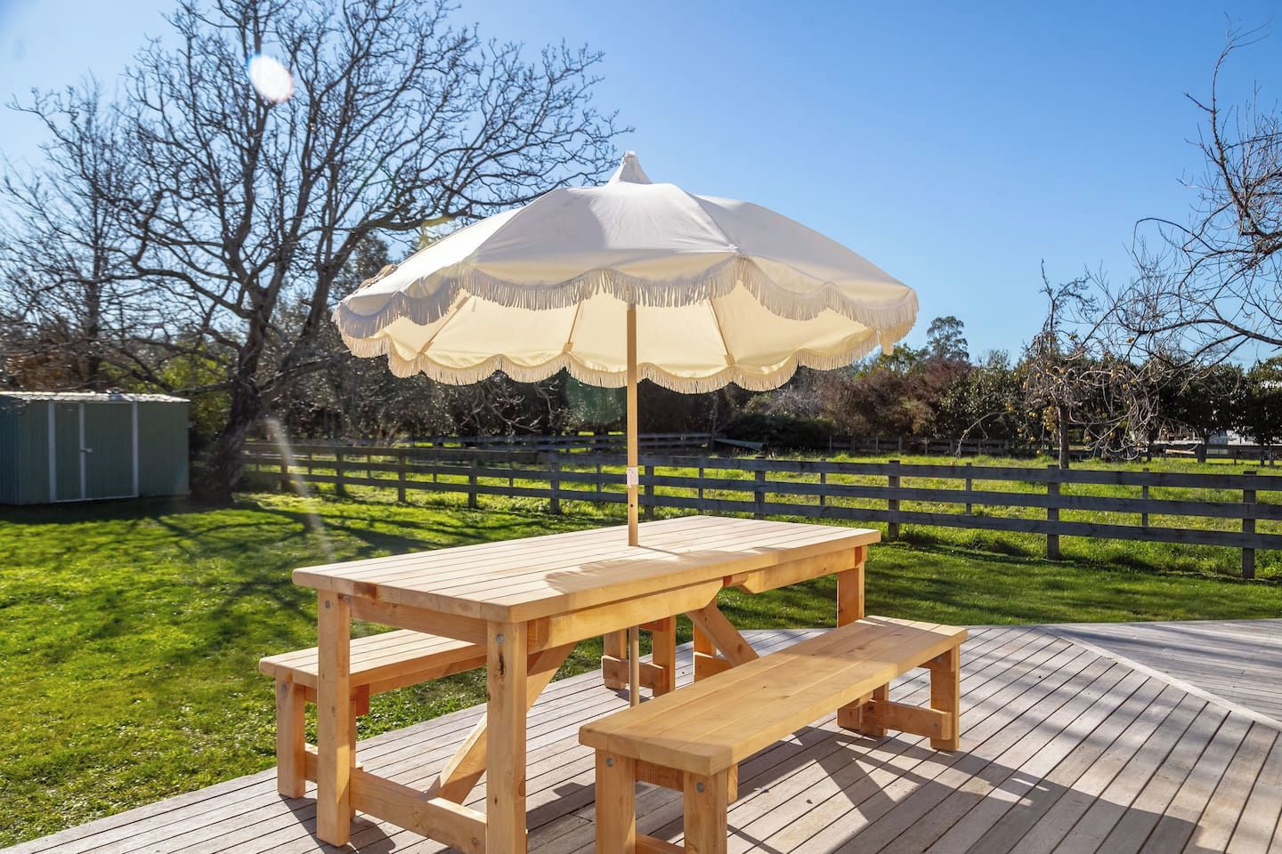 En træterrasse med et picnicbord og bænk, en parasol og en indhegnet have med træer.