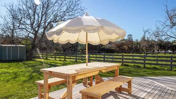 En træterrasse med et picnicbord og bænk, en parasol og en indhegnet have med træer.