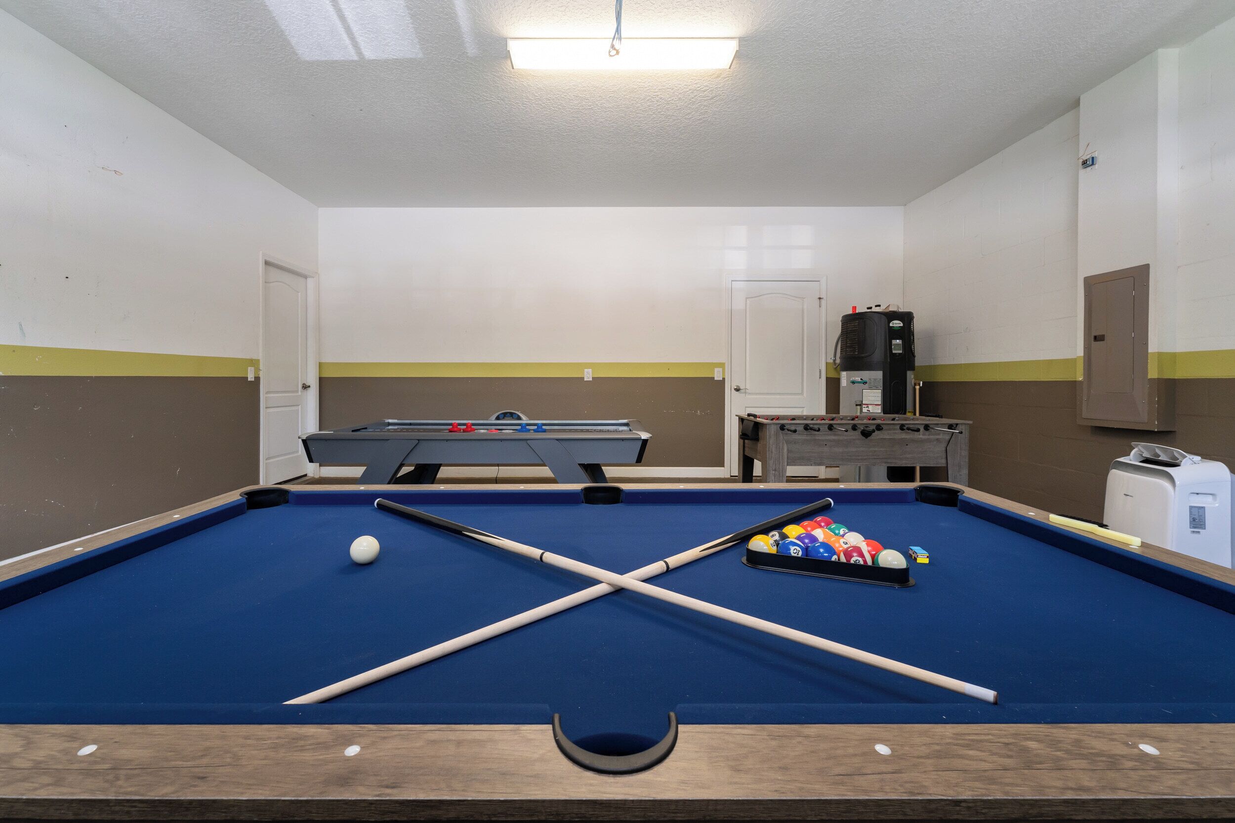 A pool table with a blue felt surface, pool cues, and balls, a coffee machine, and a trash bin in a room with white walls and yellow stripes.