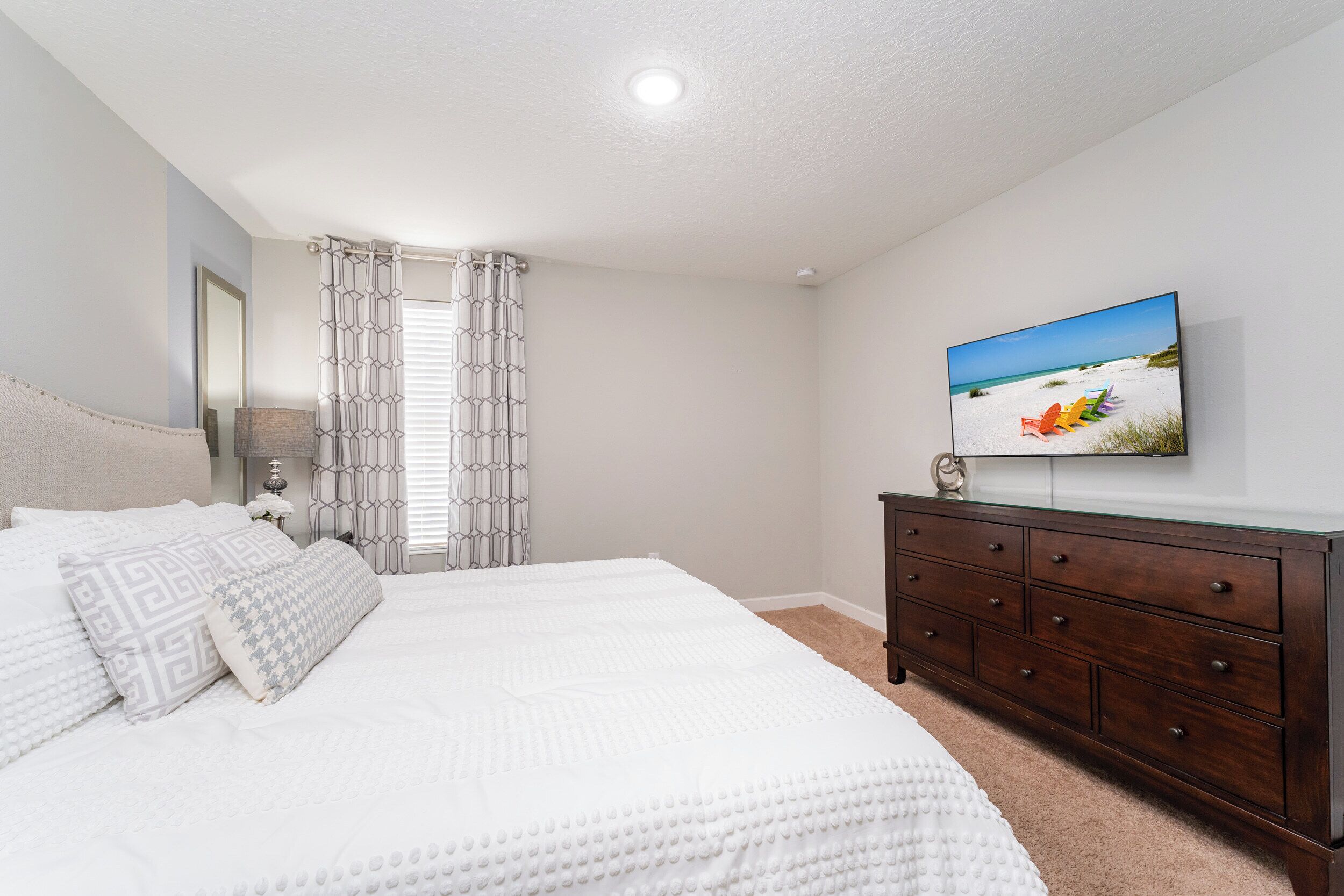 A bedroom with a large bed, a dresser, a television on the wall, and a window with curtains.