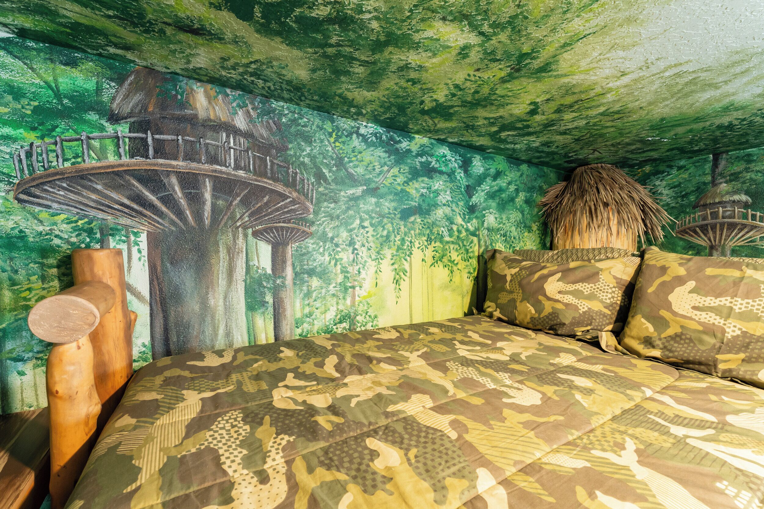 Un dormitorio con una colcha de estampado camuflado, un cabecero de madera y un mural de un bosque con una casa en el árbol.