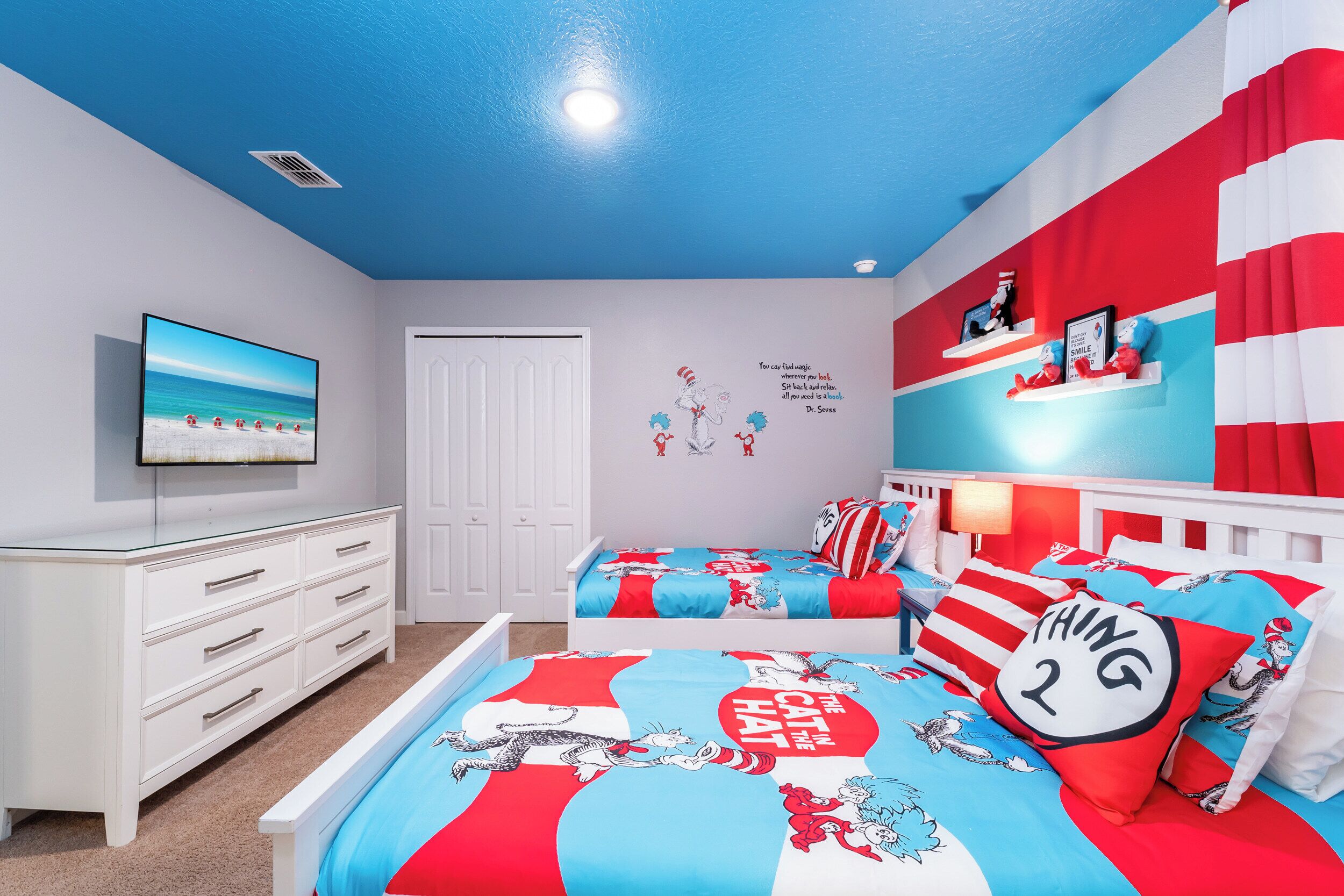 Ein Schlafzimmer mit Fernseher, weißem Schminktisch und zwei Betten mit Bettwäsche im Stil von 'The Cat in the Hat' sowie einem roten und weißen Streifenvorhang.