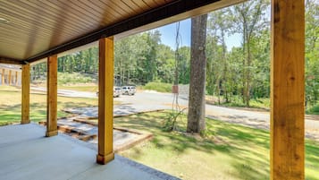 Une véranda couverte avec des poutres en bois et un sol en béton, donnant sur une zone verdoyante avec des arbres et un véhicule garé.