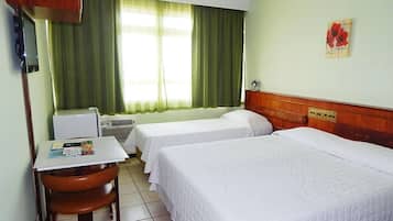 Habitación de hotel con dos camas individuales, un escritorio pequeño con una silla, un radiador y una ventana con cortinas verdes.