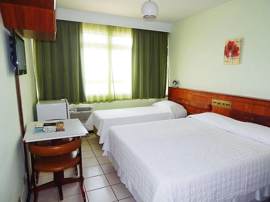 Habitación de hotel con dos camas individuales, un escritorio pequeño con una silla, un radiador y una ventana con cortinas verdes.