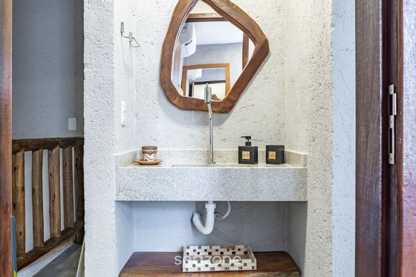 Casa de banho com parede branca texturizada, espelho com moldura de madeira, lavatório branco com torneira e dois dispensadores de sabonete pretos.