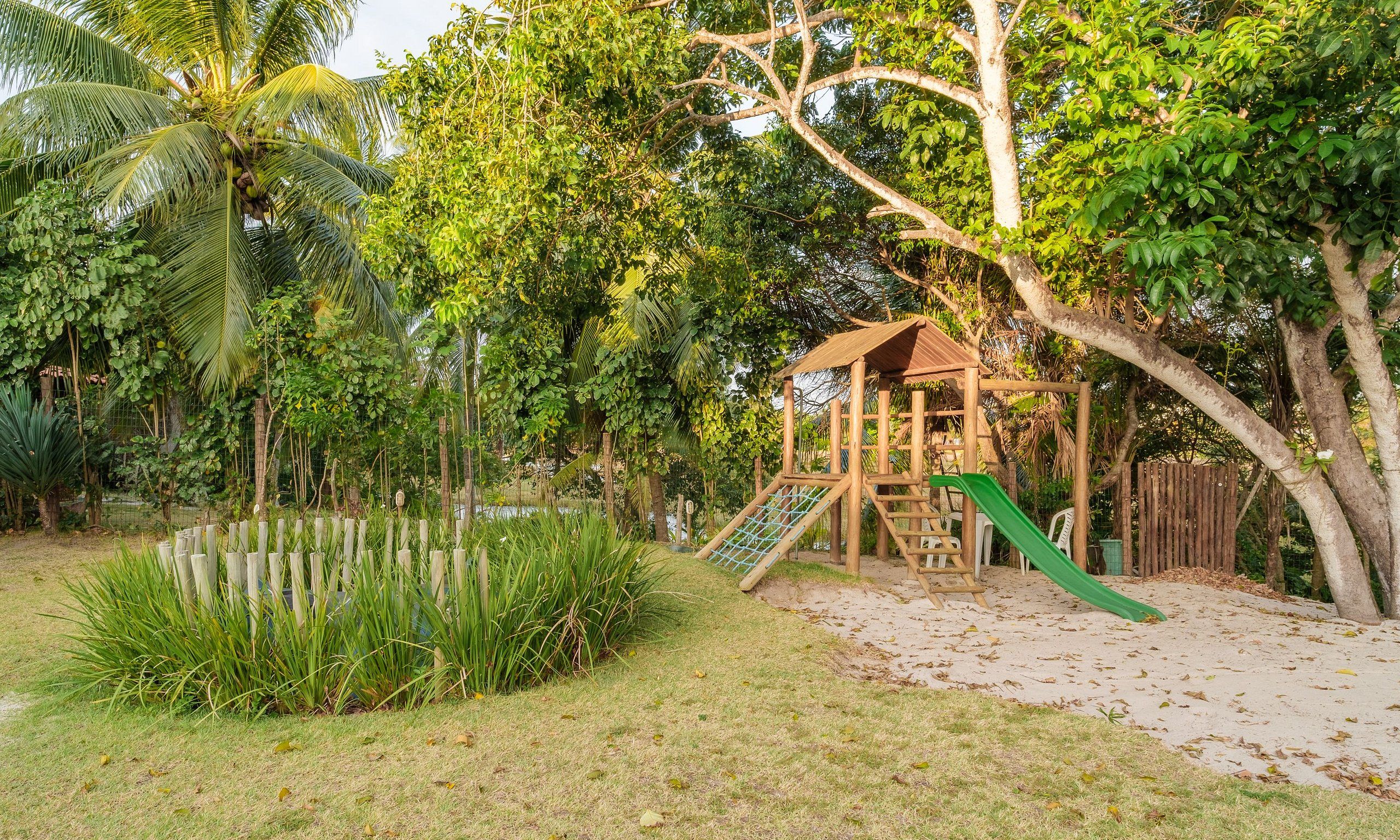 Parque infantil com escorrega e estrutura de escalada, cercado por vegetação exuberante e um lago.