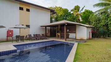 Uma piscina com deck revestido, área de estar coberta ao ar livre e um edifício com telhado telhado.