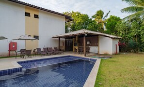 Una piscina con solárium de baldosas, una zona de estar exterior cubierta y un edificio con tejado de cerámica.