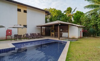 Uma piscina com deck revestido, área de estar coberta ao ar livre e um edifício com telhado telhado.