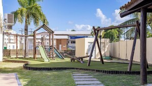 A playground with slides and swings, surrounded by a fenced area with a grassy lawn and a paved walkway. - Condominium With a Pool 850m From Praia do Sul, Ilheus/ba VGT (Ilhéus)