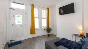 Una sala de estar con un sofá gris, una mesita con una lámpara, un televisor en la pared y una puerta con cortinas amarillas.