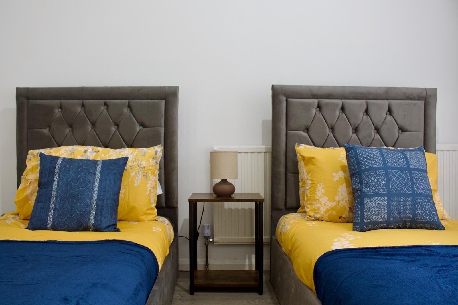 Una cama doble con cabecera gris tapizada, una mesita de noche pequeña con lámpara, y ropa de cama floral amarilla y azul a juego.