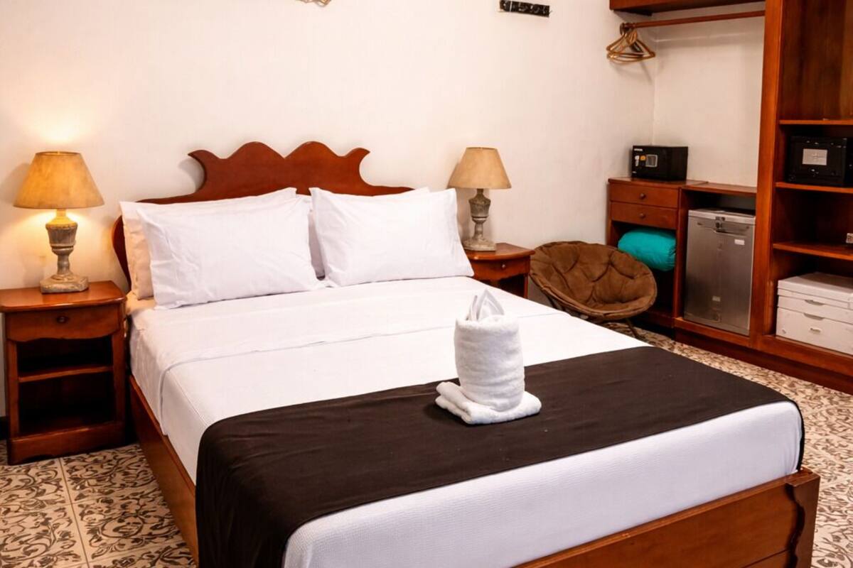 A neatly made bed with white linens and a brown headboard, two bedside lamps, a small bedside table, a wooden nightstand, a decorative wall hanging, a wardrobe, and a tiled floor with a floral pattern.