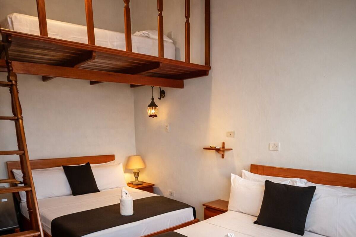 A hotel room with two beds, a wooden ladder, a patterned floor, and a small table with a lamp.