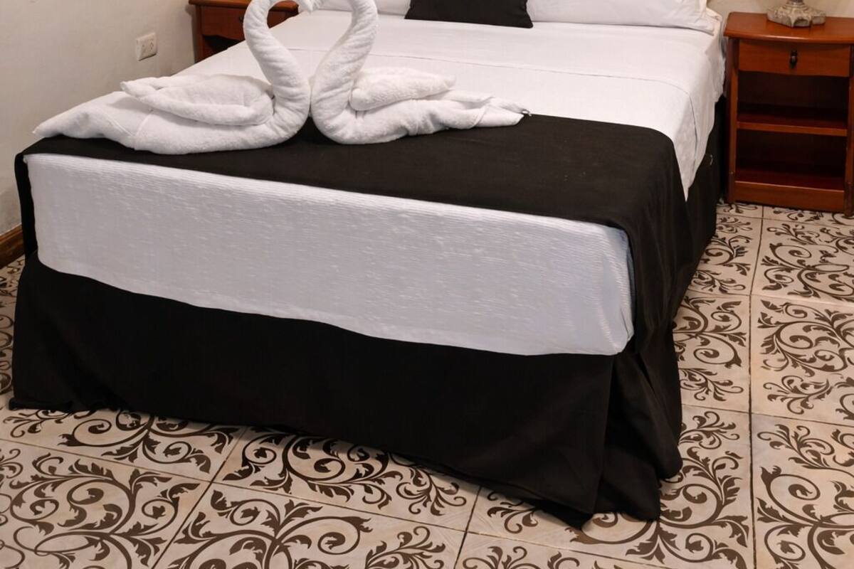 A neatly made bed with a white and black bedspread, two pillows, and a white towel folded on top. There are two bedside tables with lamps, a decorative wall hanging above the bed, and a patterned floor.