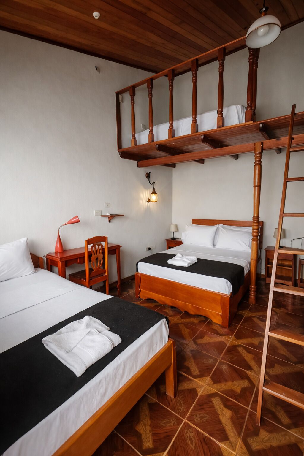 A hotel room with two beds, a wooden ceiling, a staircase, and a wall-mounted light fixture.