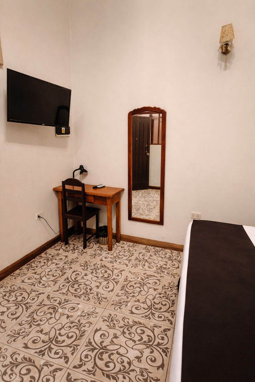 A bedroom with a bed, a desk with a chair, a TV mounted on the wall, a mirror, and patterned floor tiles.