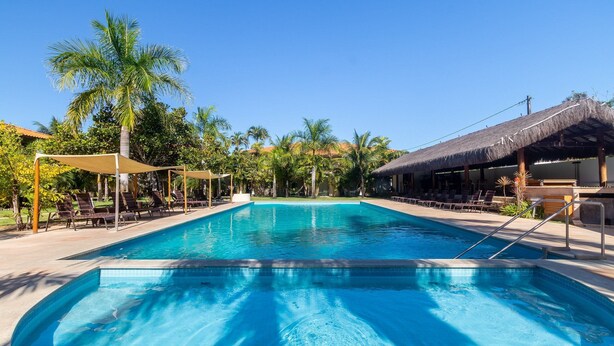 Una piscina con sillas de descanso y sombrillas, rodeada de palmeras y una estructura de techo de paja.