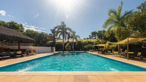 Una piscina con sillones de descanso y sombrillas, rodeada de palmeras y un edificio con techo de paja.