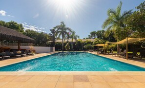 Una piscina con sedie a sdraio e ombrelloni, circondata da palme e un edificio con tetto di paglia.