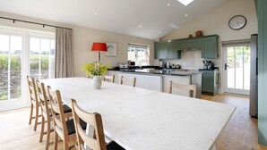 A spacious kitchen with a large island, a dining area with a white table and wooden chairs, and a view of the outdoors through large windows.