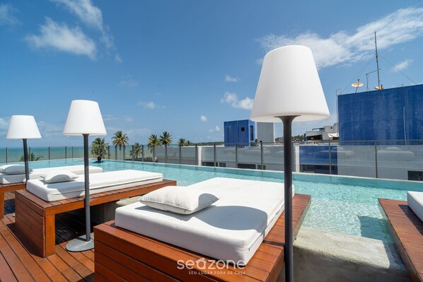A rooftop pool area with lounge chairs and a large umbrella, overlooking a clear blue sky and ocean.
