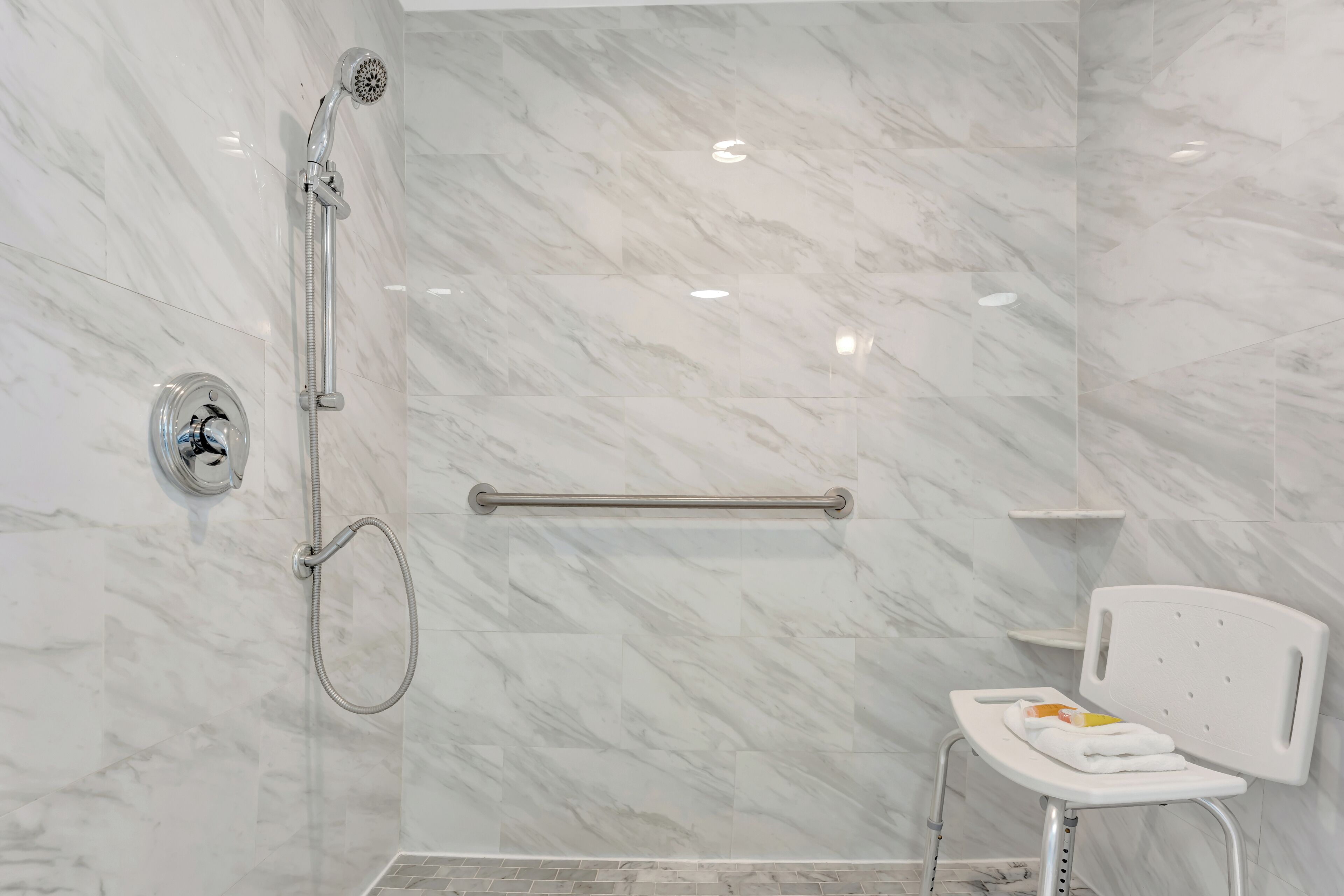 Baño con ducha, barra de apoyo y una silla blanca con toalla.