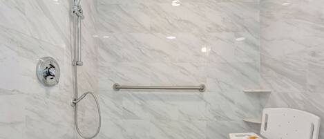 Baño con ducha, barra de apoyo y una silla blanca con toalla.