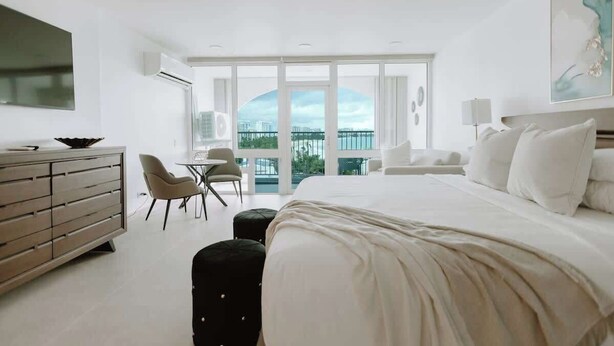 A modern hotel room with a large bed, a desk, a chair, and a balcony with a view.